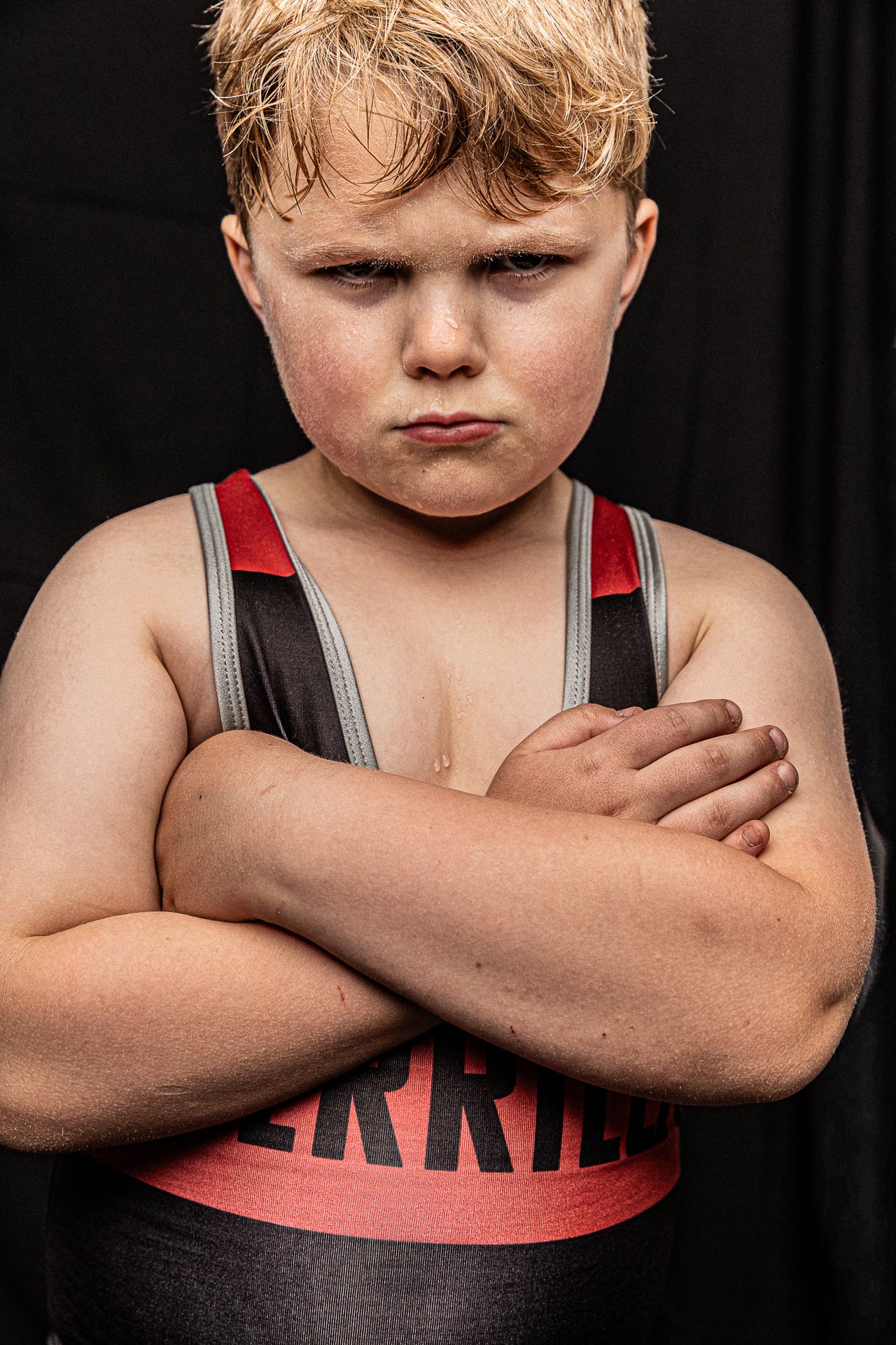 Young wrestler in black and red singlet stands with arms crossed, displaying a determined expression against dark background.