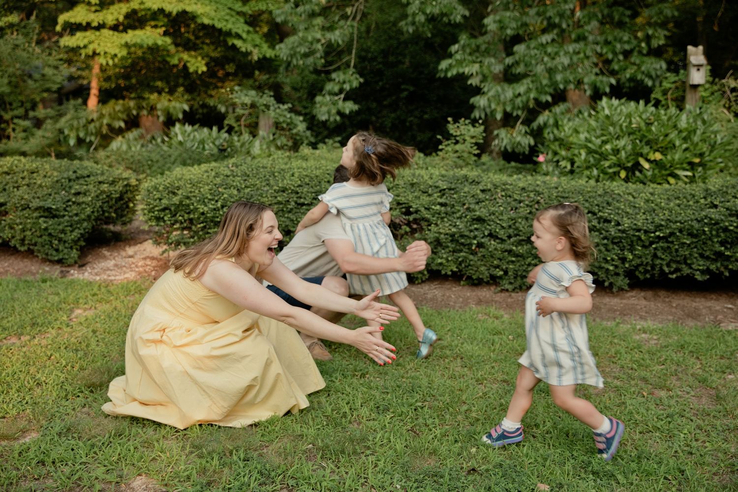 A joyful mother in a yellow dress reaches for her two young daughters running toward her outdoors.