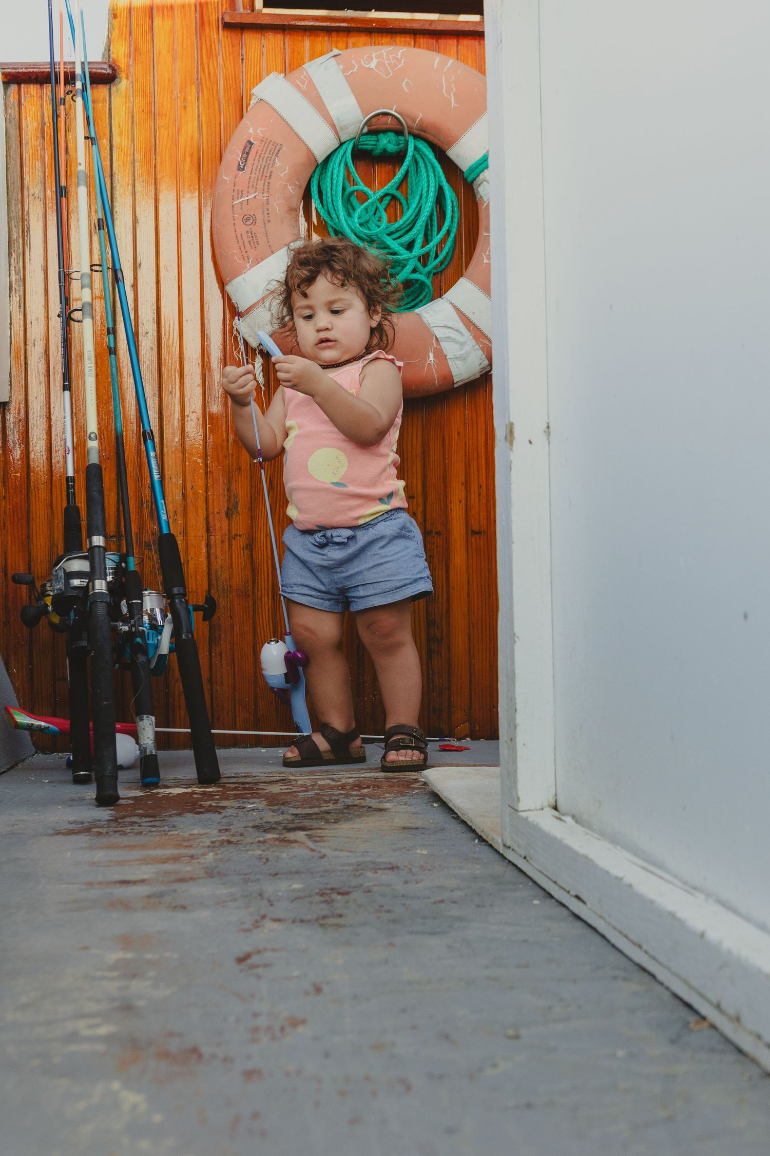 Young toddler girl standing on boat deck near fishing rods and life preserver ring on wooden wall.