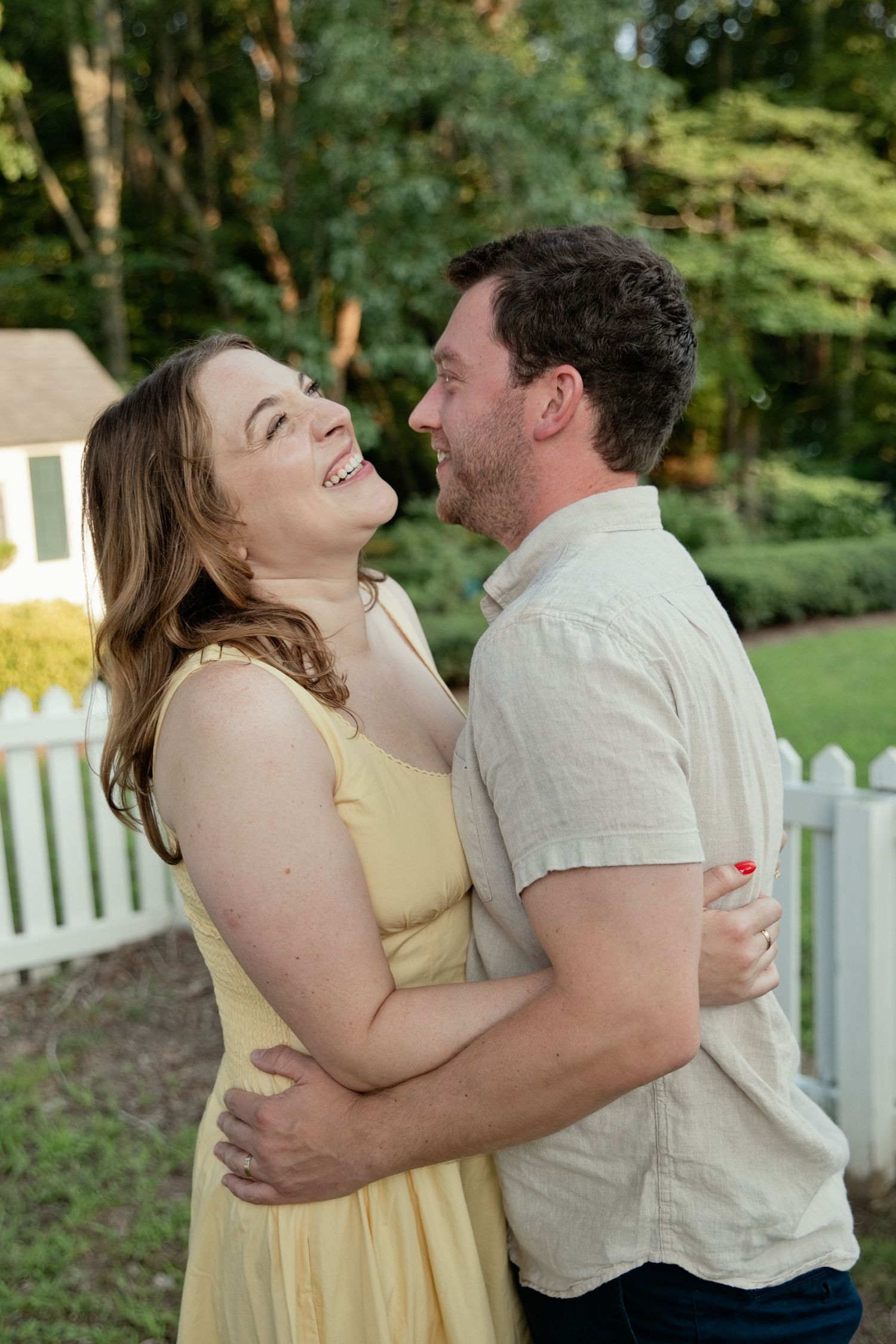 A laughing couple embraces outdoors near a white picket fence surrounded by lush green trees.