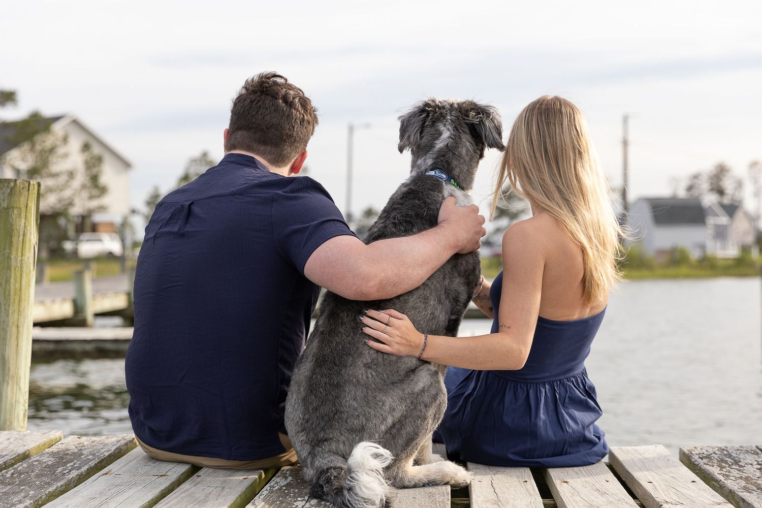 Couple sitting on a wooden dock with their gray schnauzer dog, overlooking a calm waterway on a cloudy day.