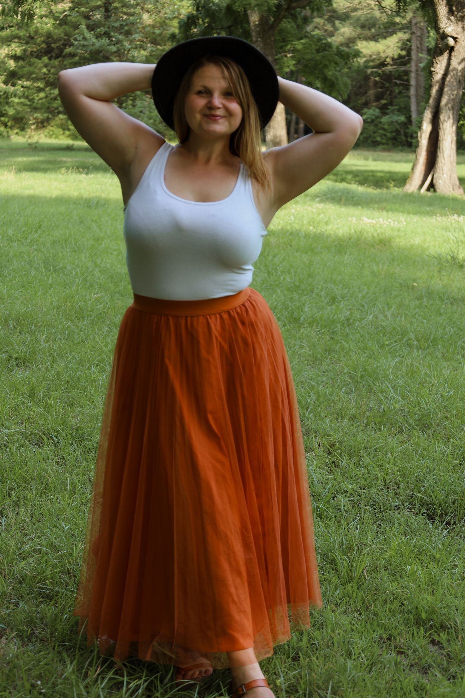 Woman in white tank top, rust orange tulle skirt, and black hat posing outdoors on green grass.
