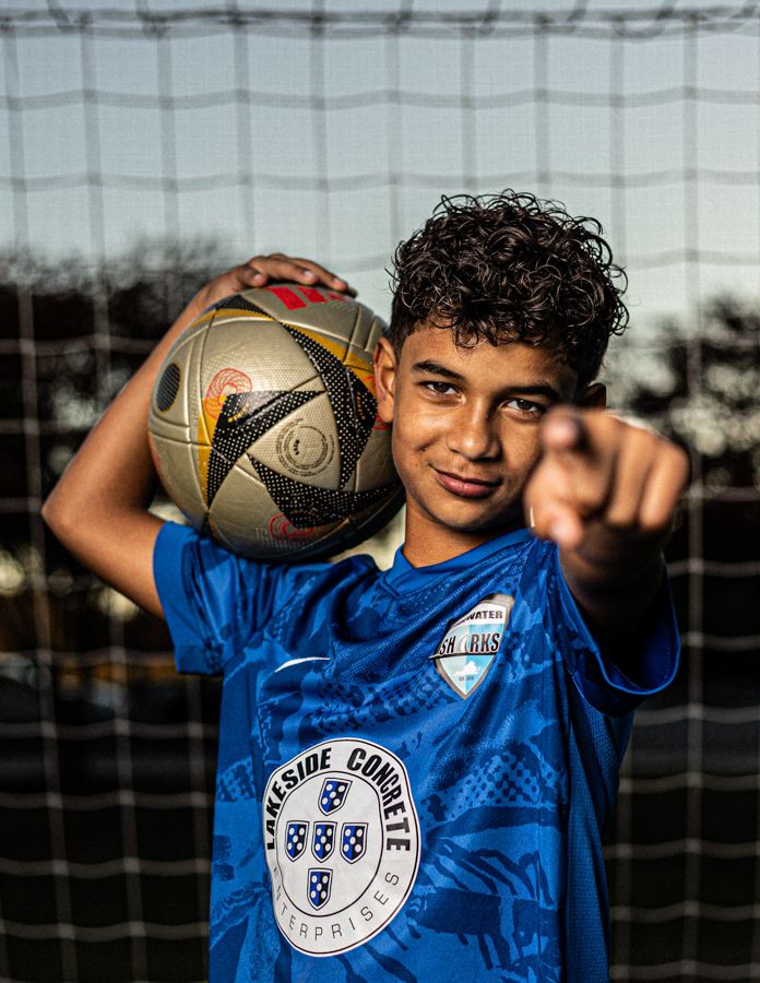 Young soccer player in blue jersey holding ball, pointing at camera in front of goal net.