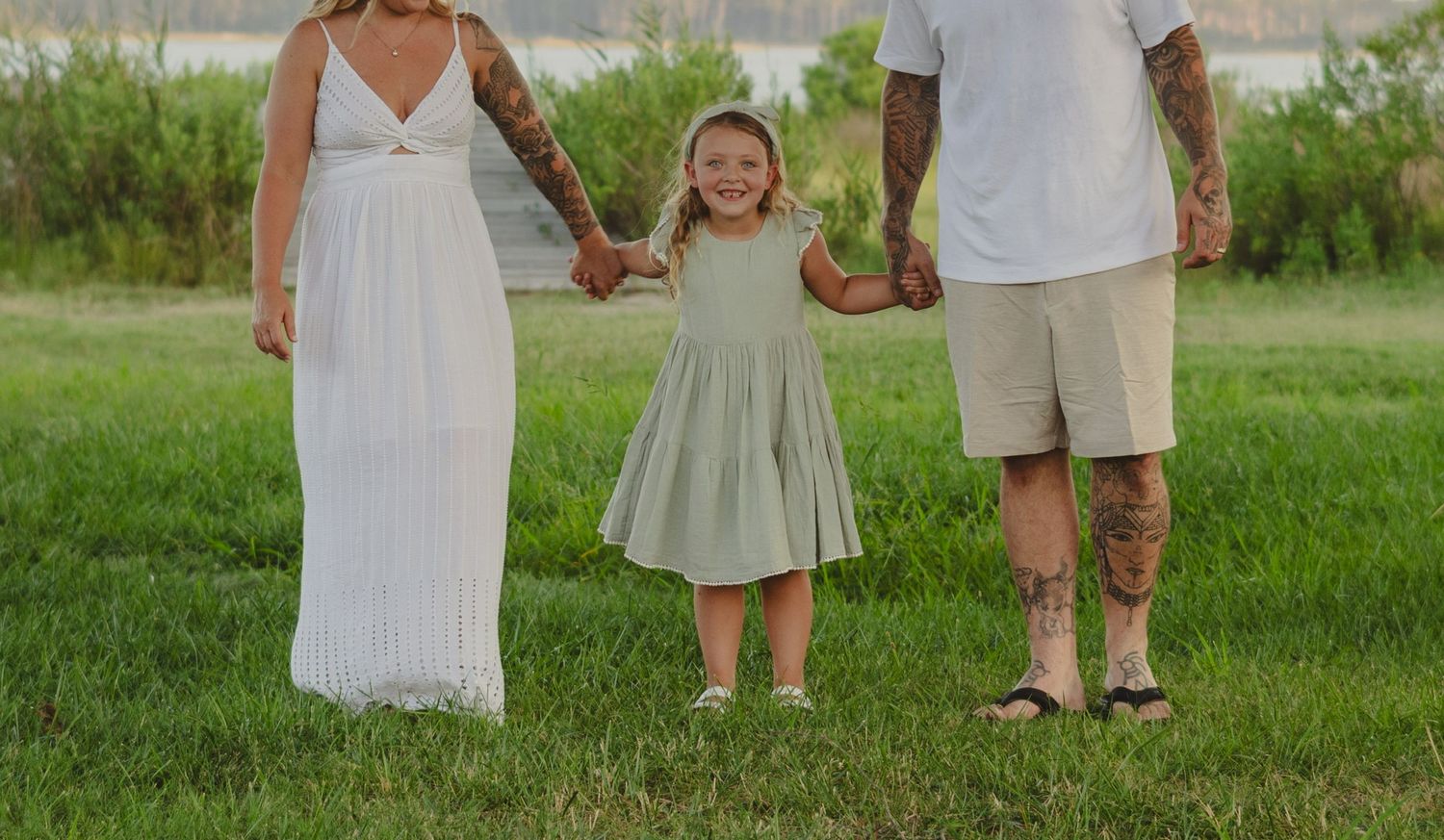 Family of three holding hands outdoors, parents with tattoos and young daughter in sage green dress on grass.
