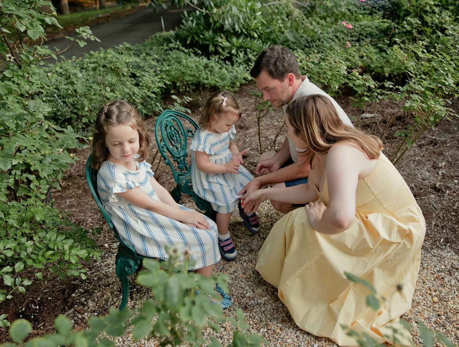 Family of four enjoying a garden moment, parents and two girls in pastel dresses among lush greenery.