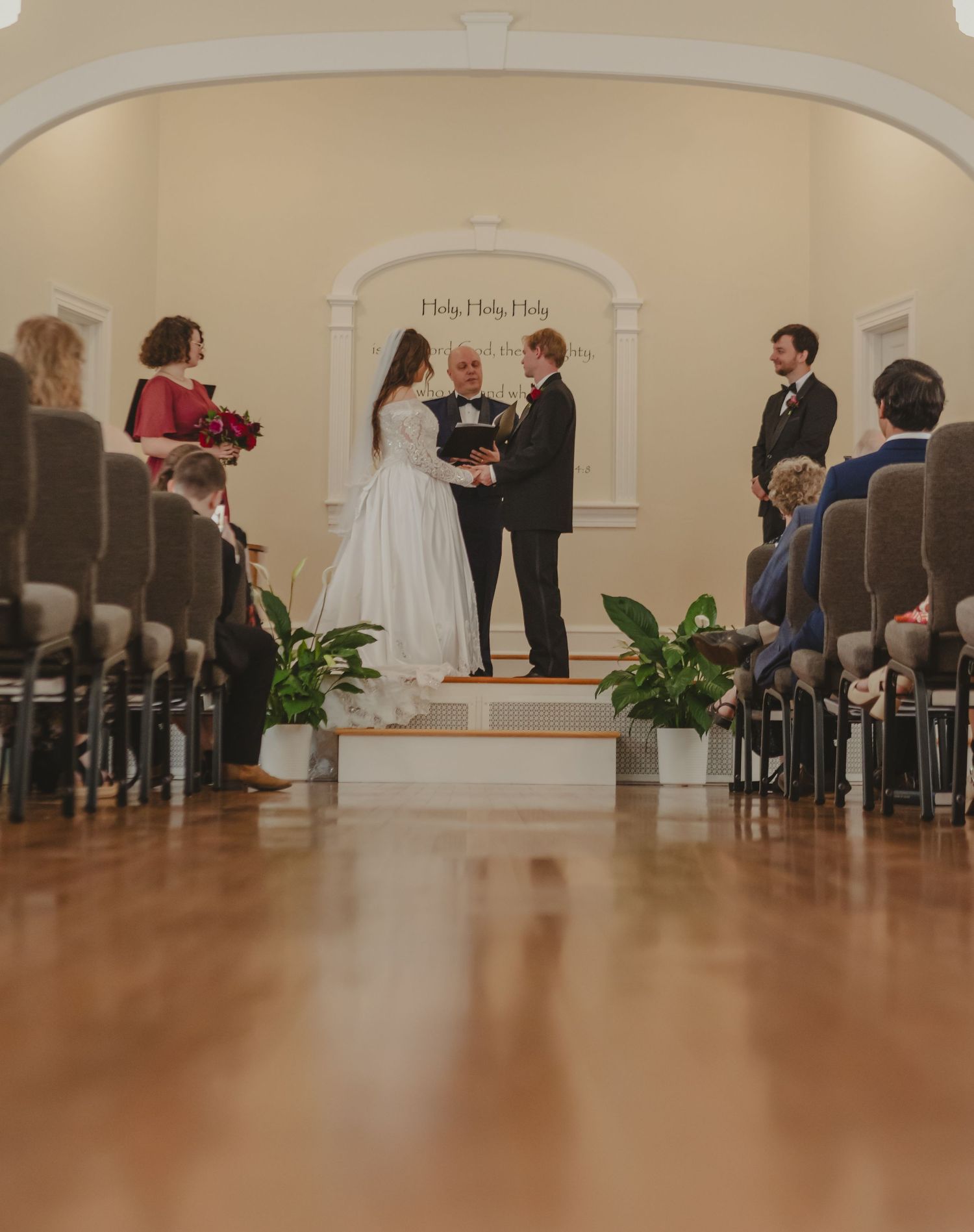 A bride and groom exchange vows at a chapel altar, surrounded by seated guests in a warmly lit wedding ceremony.