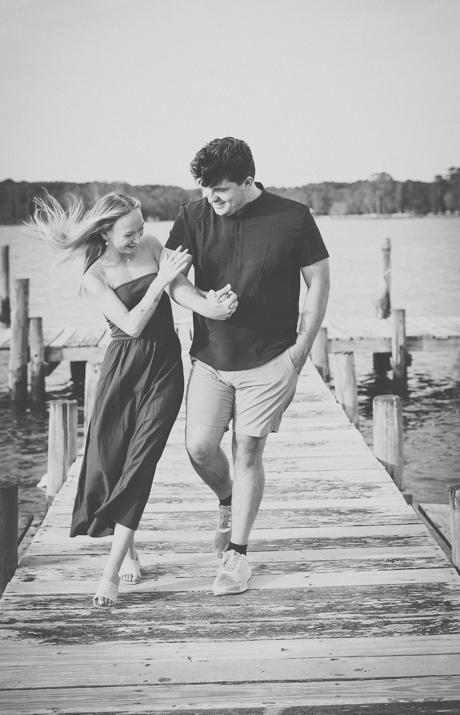 Couple laughing and walking together on wooden pier by lake in black and white photo.