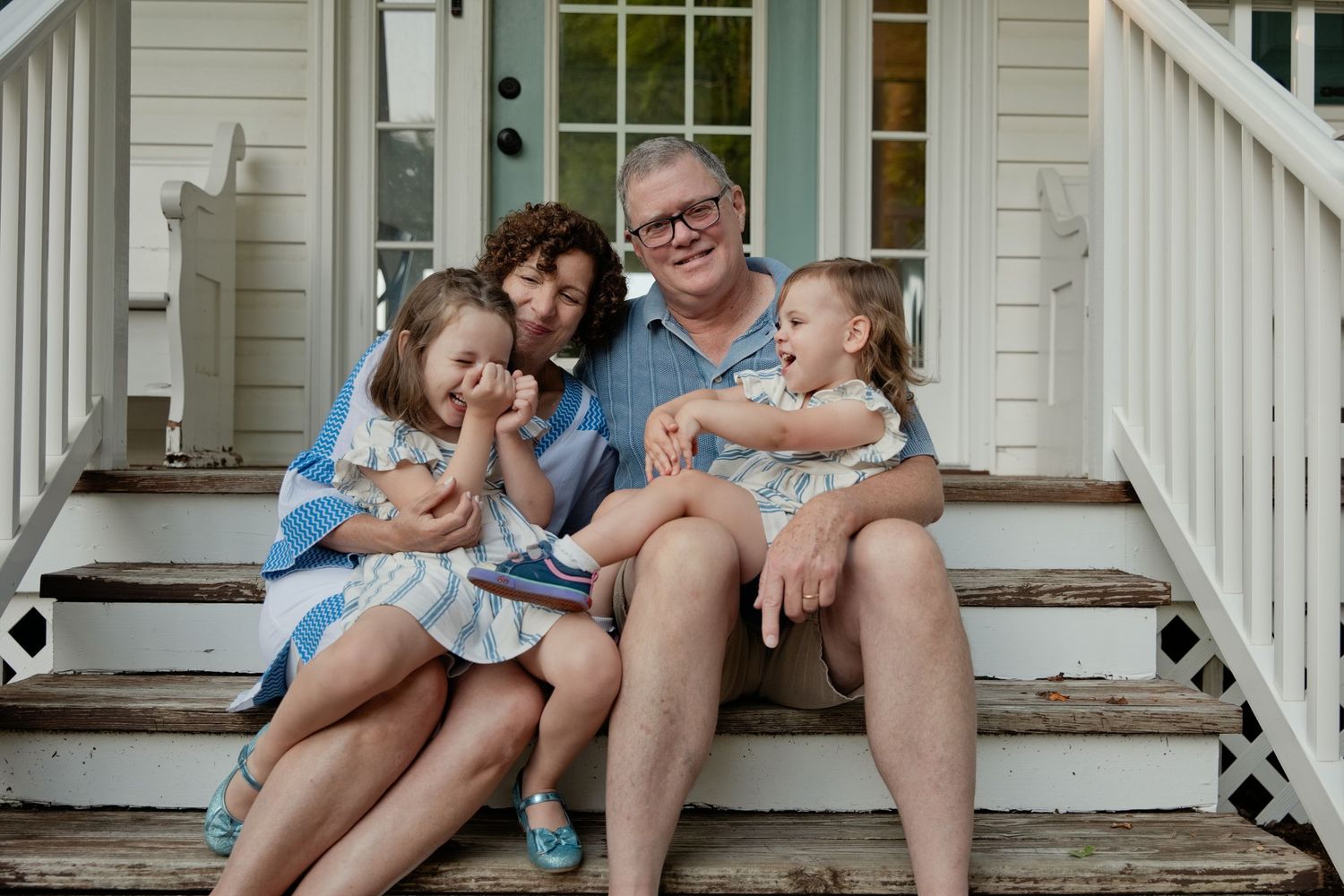 Family of four sitting on porch steps, grandparents with two young girls laughing together.