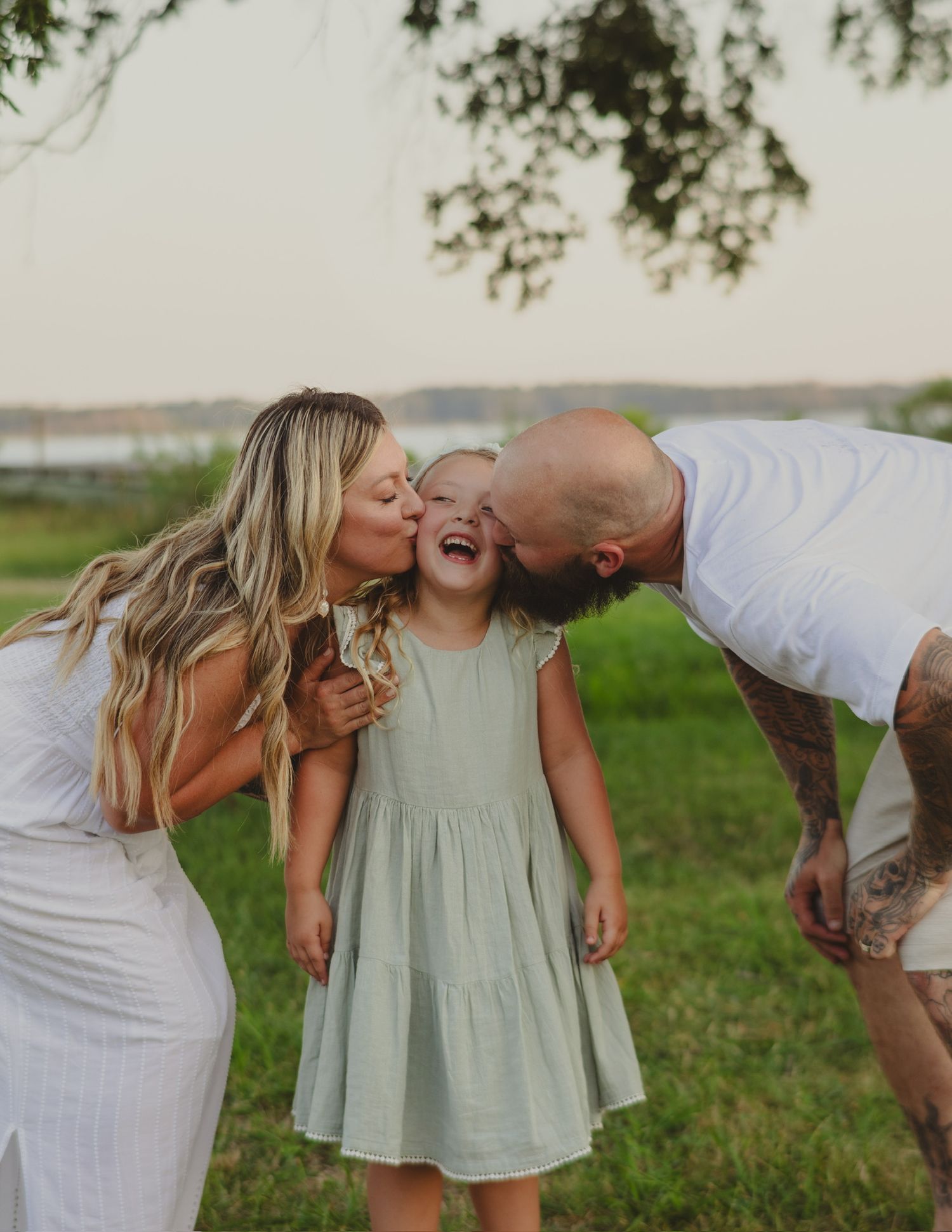 Parents kissing their laughing little girl on the cheeks outdoors in a green field during a family photo session.