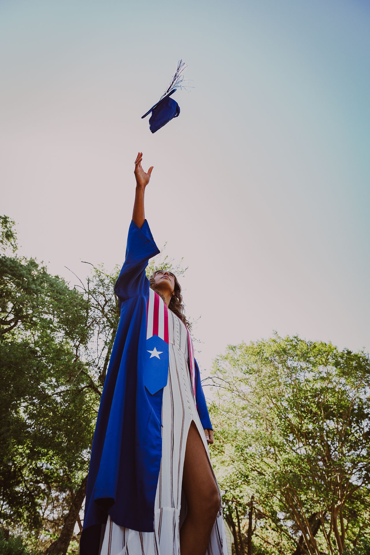 Graduate in blue cap and gown tossing mortarboard into the sky, celebrating outdoors among green trees.
