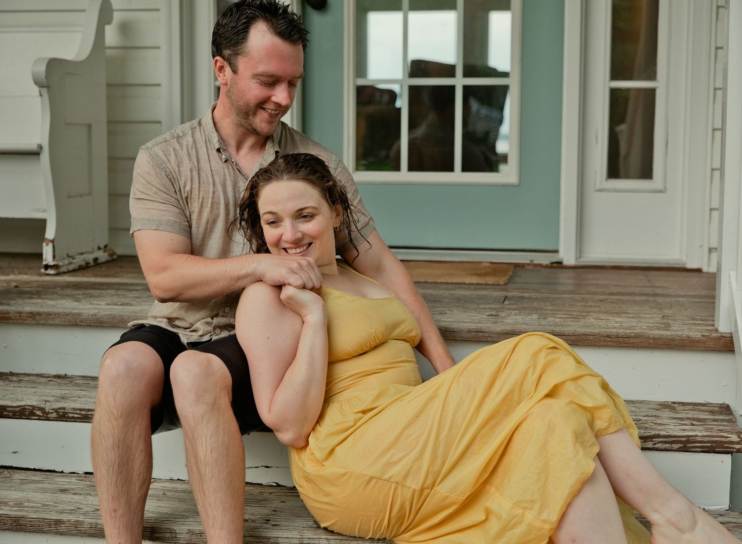 A happy couple sits together on porch steps, woman in yellow dress leaning against her partner.