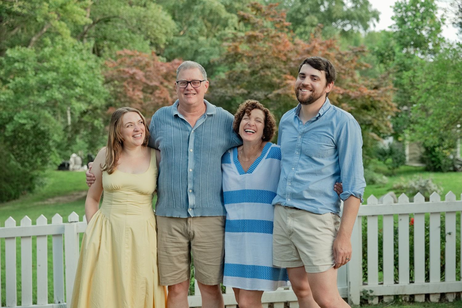 Happy family of four smiling together outdoors near a white picket fence surrounded by lush green trees.