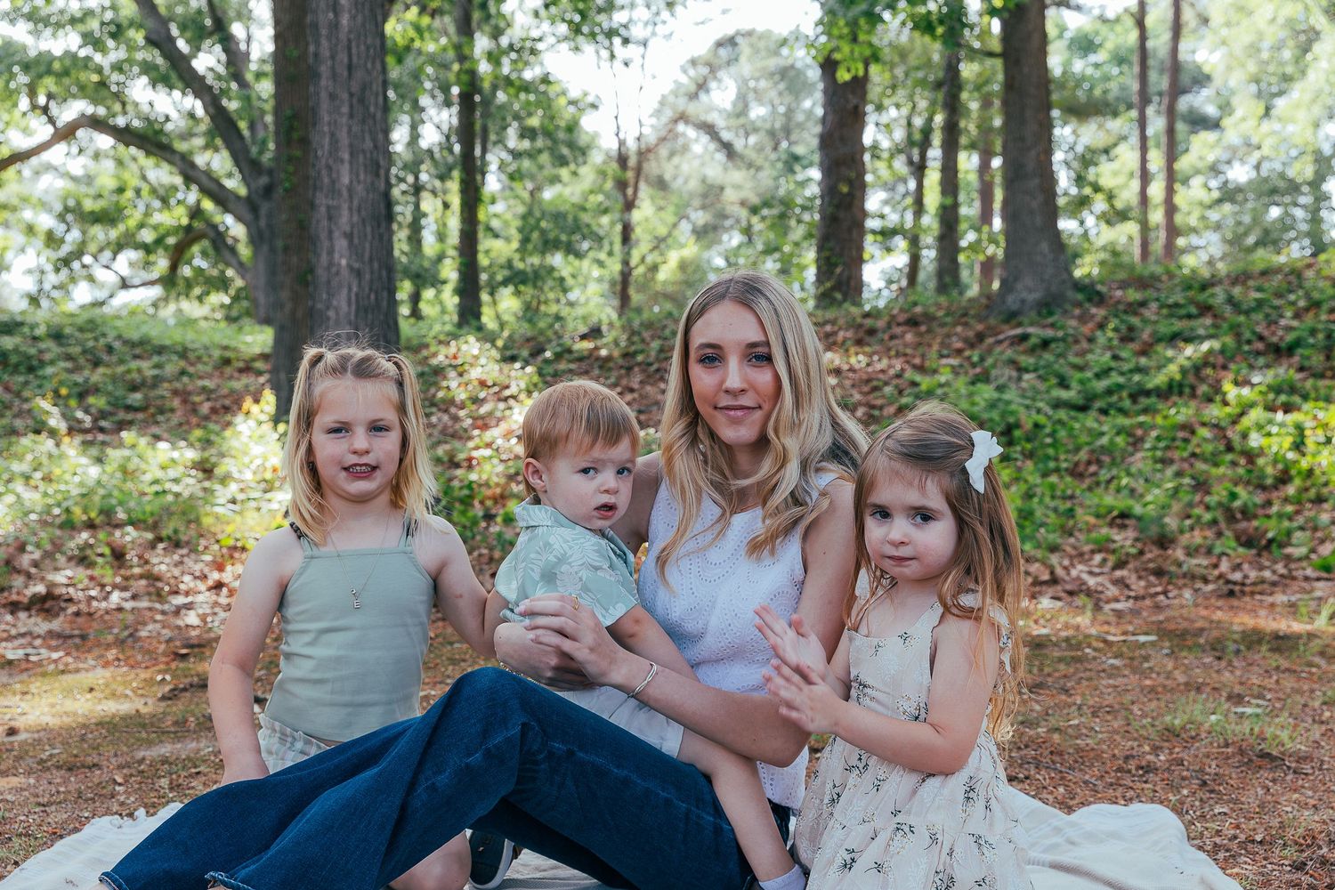 A blonde woman sits outdoors in a wooded park with three young children on a white blanket for a family portrait.