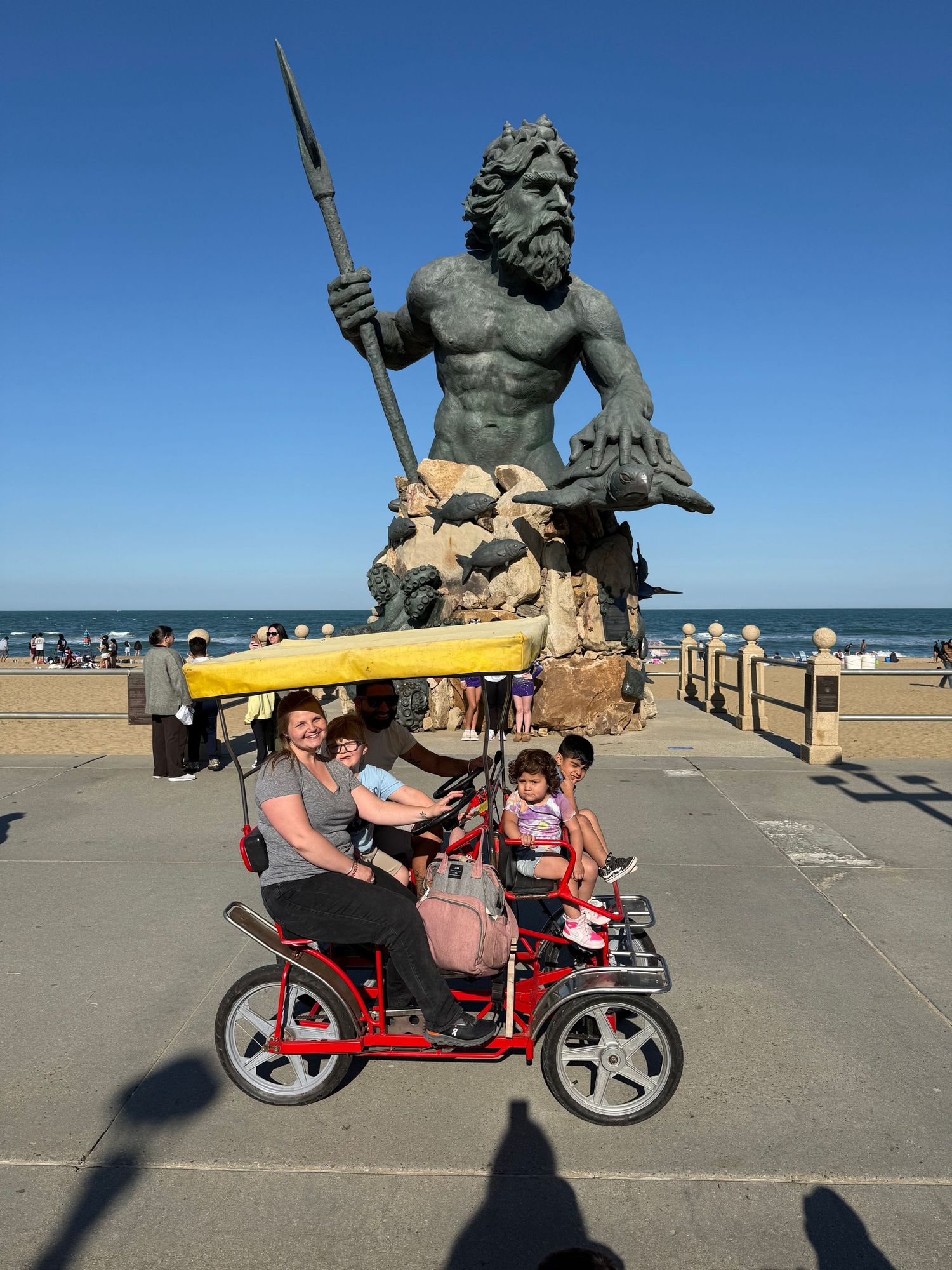 Family riding a red surrey bike near the King Neptune statue at Virginia Beach boardwalk on a sunny day.