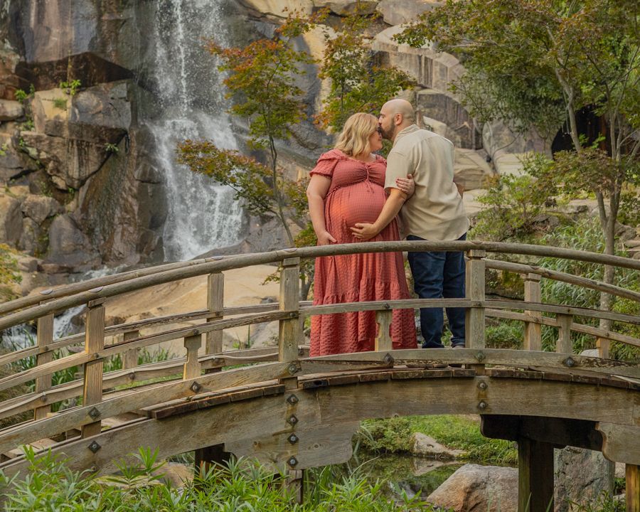 Couple sharing tender kiss on wooden bridge with waterfall backdrop during outdoor maternity photo session.
