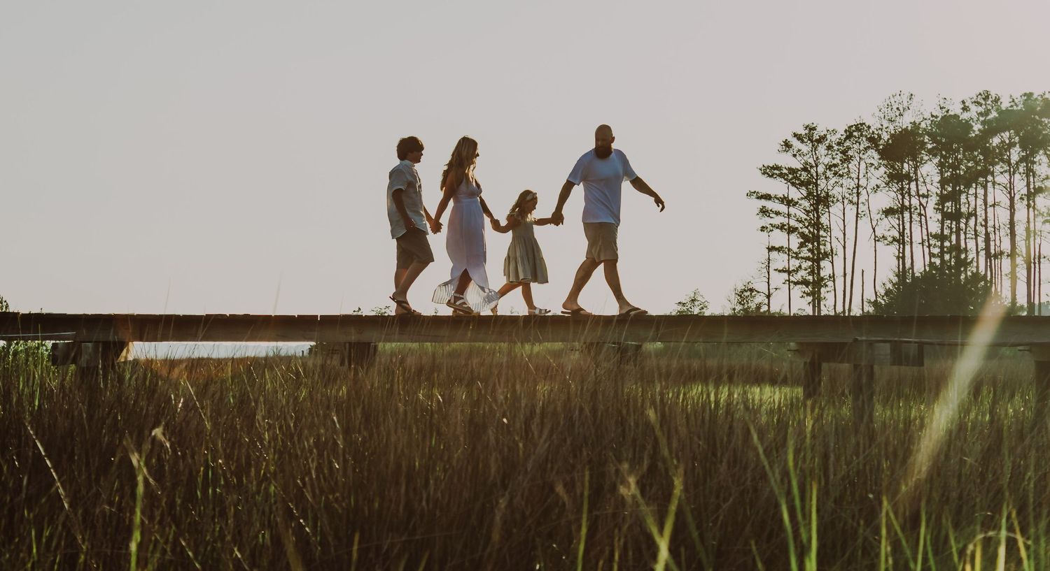 family on a pier holding hands with the sun in the background and the river grass showing