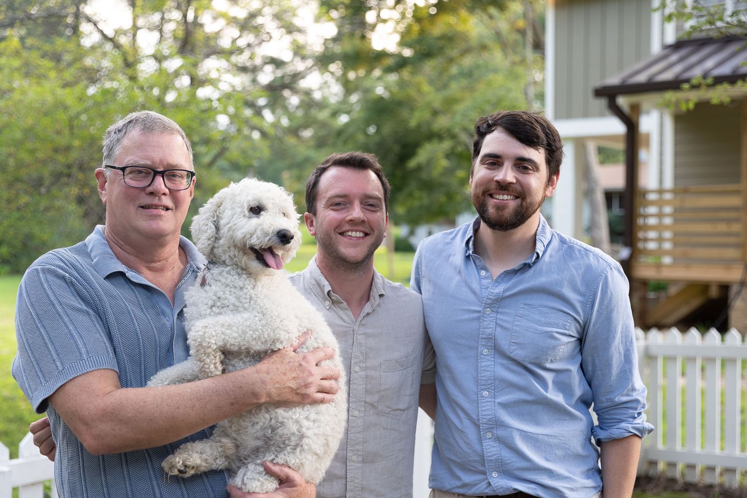 Three smiling men pose outdoors with a fluffy white dog near a white picket fence.