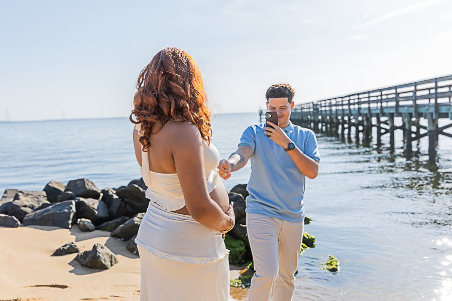A man photographs a woman in a white dress on a sunny beach near a wooden pier and rocky shoreline.