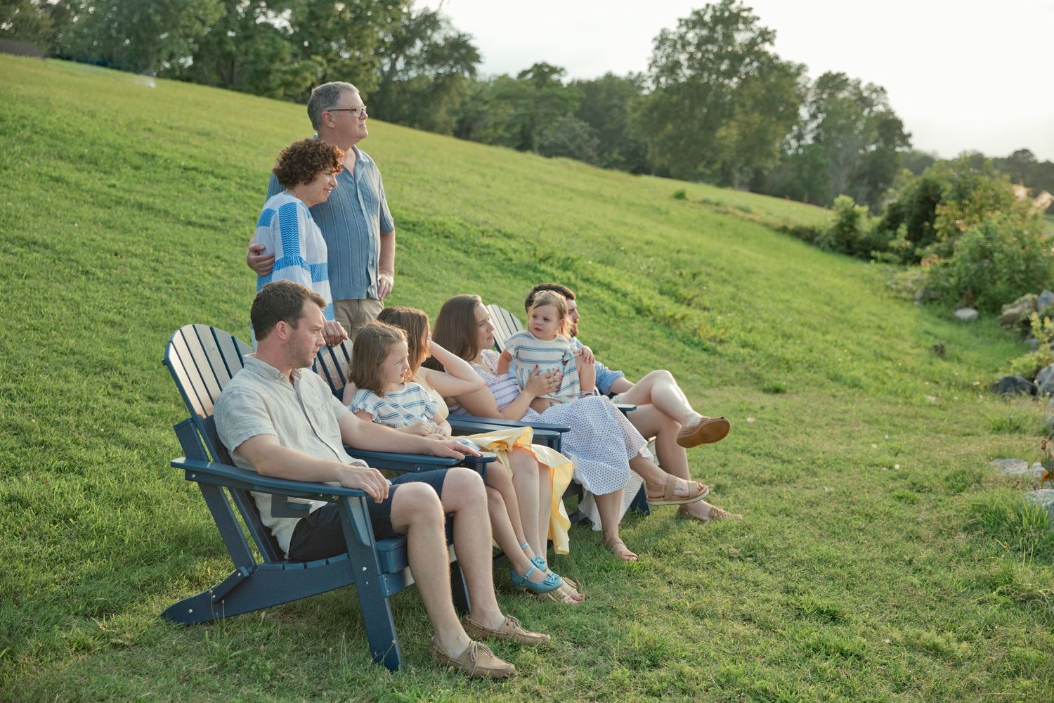 A multigenerational family relaxes together in Adirondack chairs on a sunny green hillside.