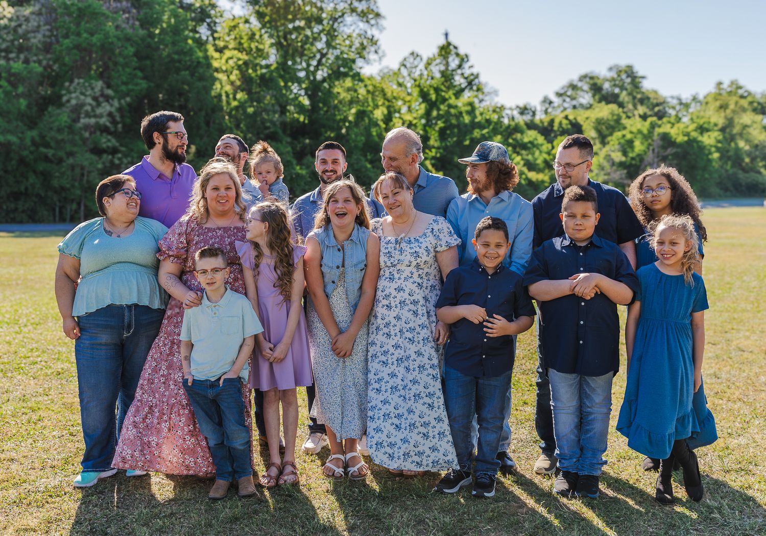 Large family group portrait outdoors on green grass, featuring adults and children dressed in colorful attire, smiling together.