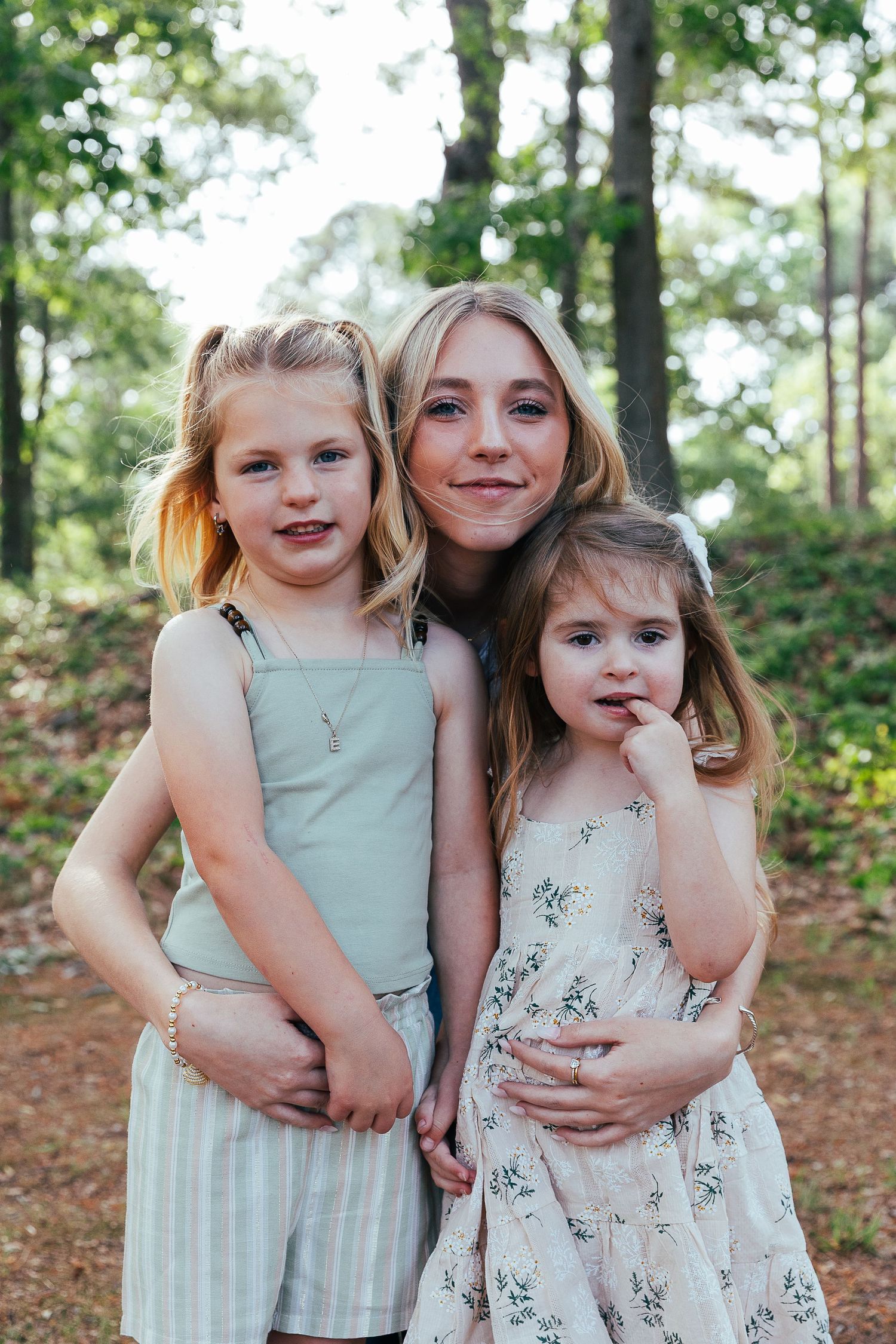 A woman hugs two young blonde girls in a sunlit forest, all wearing summer dresses and smiling at the camera.