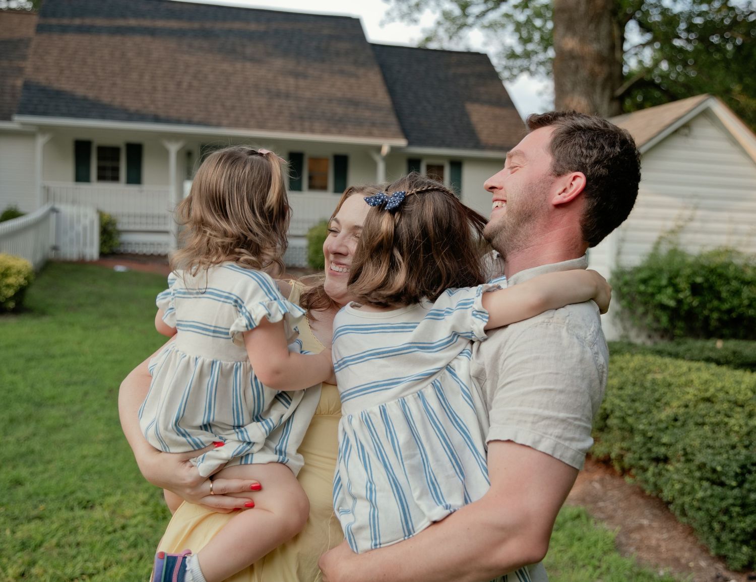 Happy family embracing outdoors in front of a white farmhouse, parents hugging two young daughters in matching striped dresses.