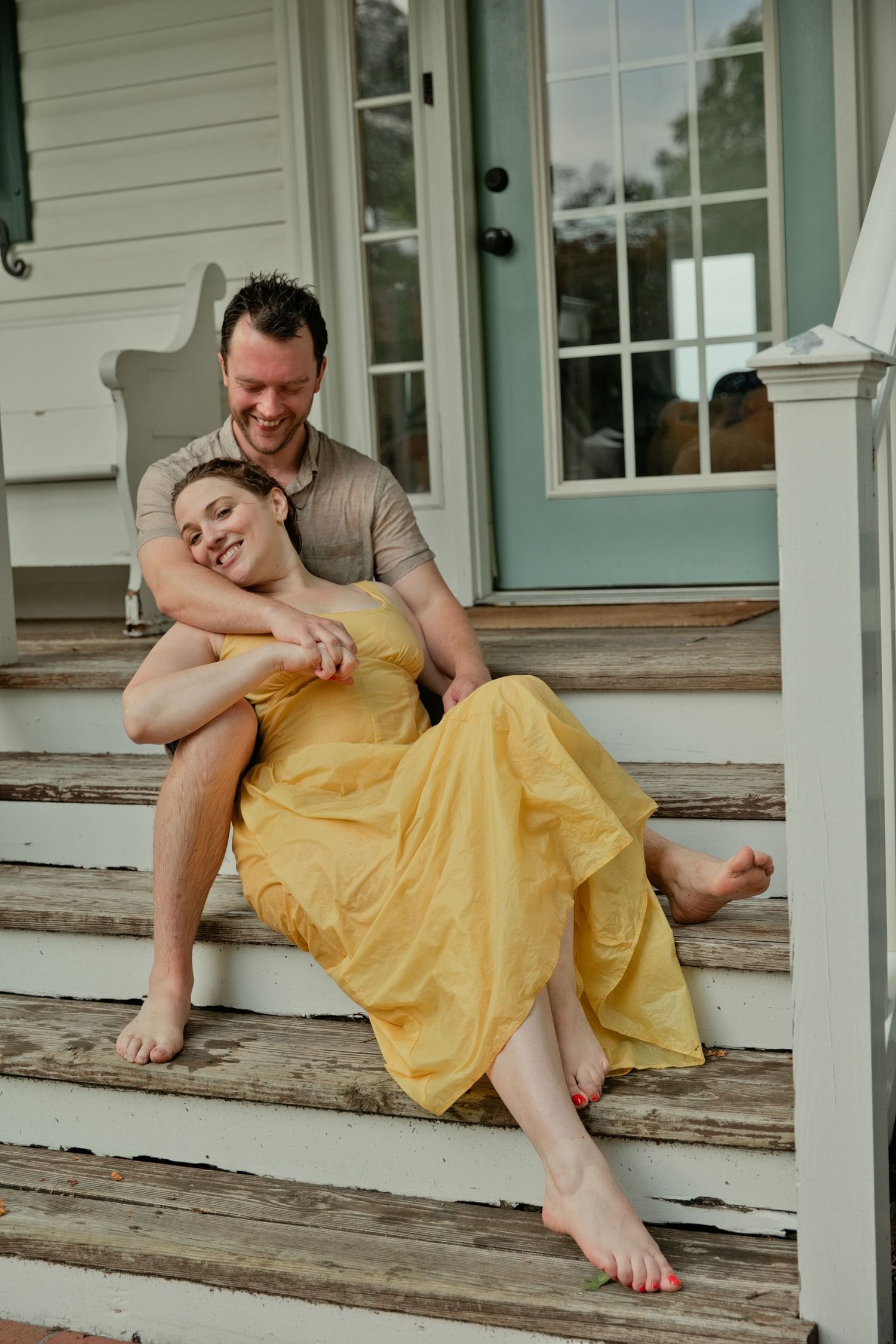 A happy barefoot couple sits closely together on rustic wooden porch steps, woman in flowing yellow dress.