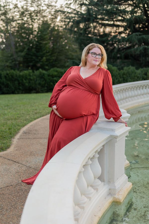 pregnant woman in a red dress leaning against a railing on a fountain of water