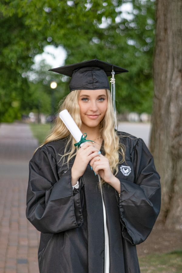 A smiling blonde woman in black graduation cap and gown holds a diploma outdoors on a tree-lined campus walkway.