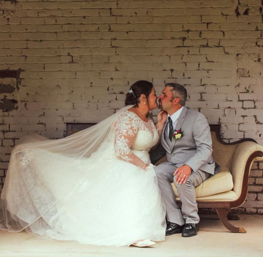 Bride and groom share a romantic kiss while seated on a vintage chaise lounge against a rustic white brick wall.