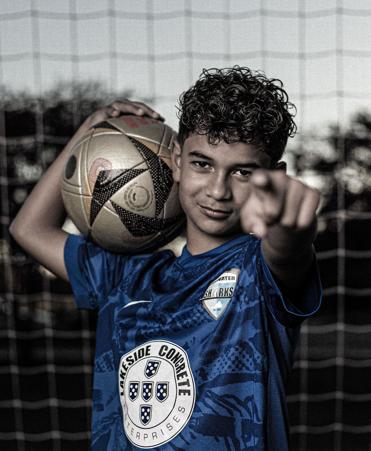 Young soccer player in blue jersey holds a ball on shoulder while pointing at camera near a goal net.