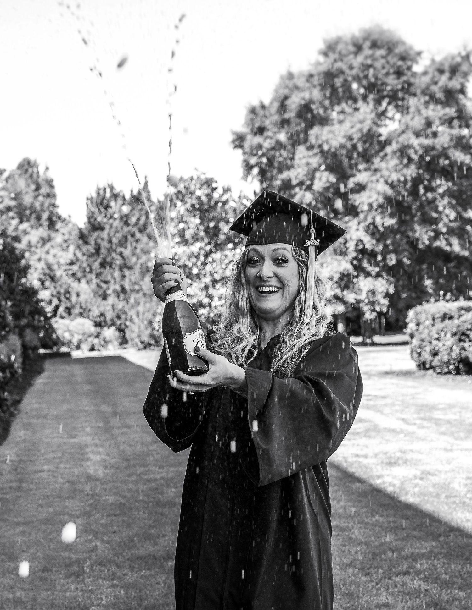 Graduate in cap and gown celebrates with champagne bottle spraying outdoors in black and white photo.