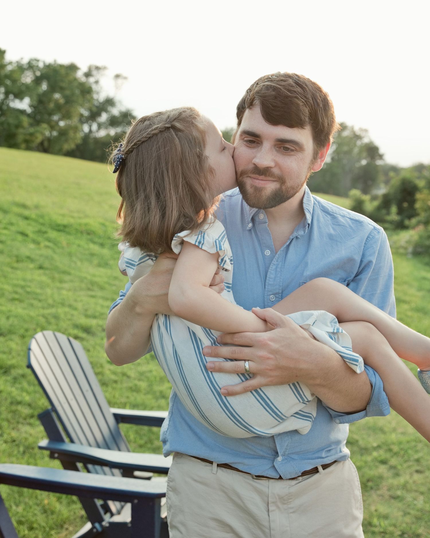 A smiling man holds a young girl who kisses his cheek in a sunny green field.