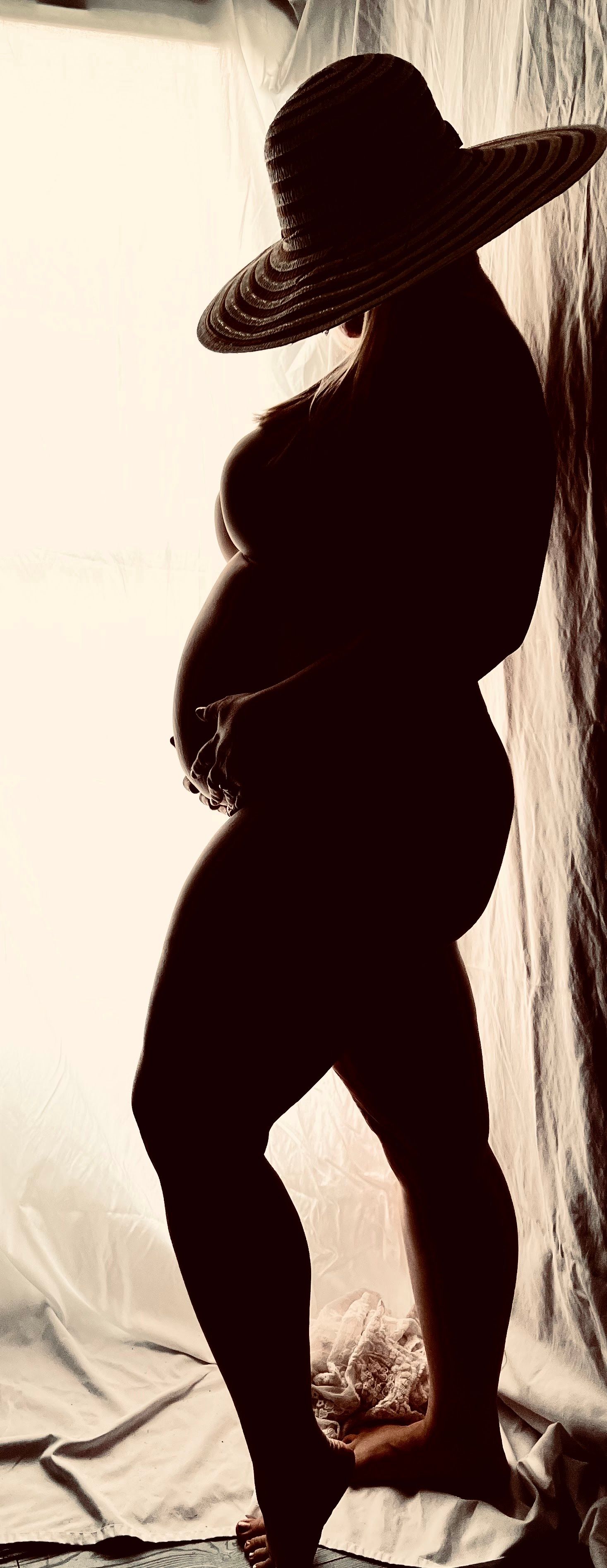 Artistic silhouette of a pregnant woman wearing a wide-brim straw hat, cradling her baby bump against a bright white backdrop.