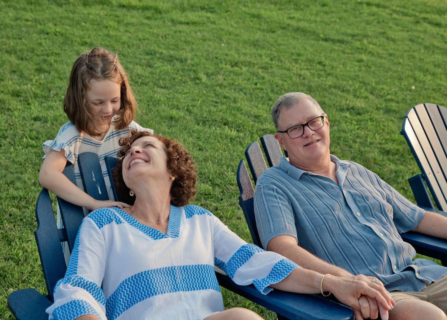 Grandparents relax in blue Adirondack chairs on green grass while a young granddaughter leans in smiling.