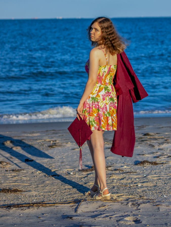 Young woman in floral dress holding graduation cap and red gown on a sunny beach.