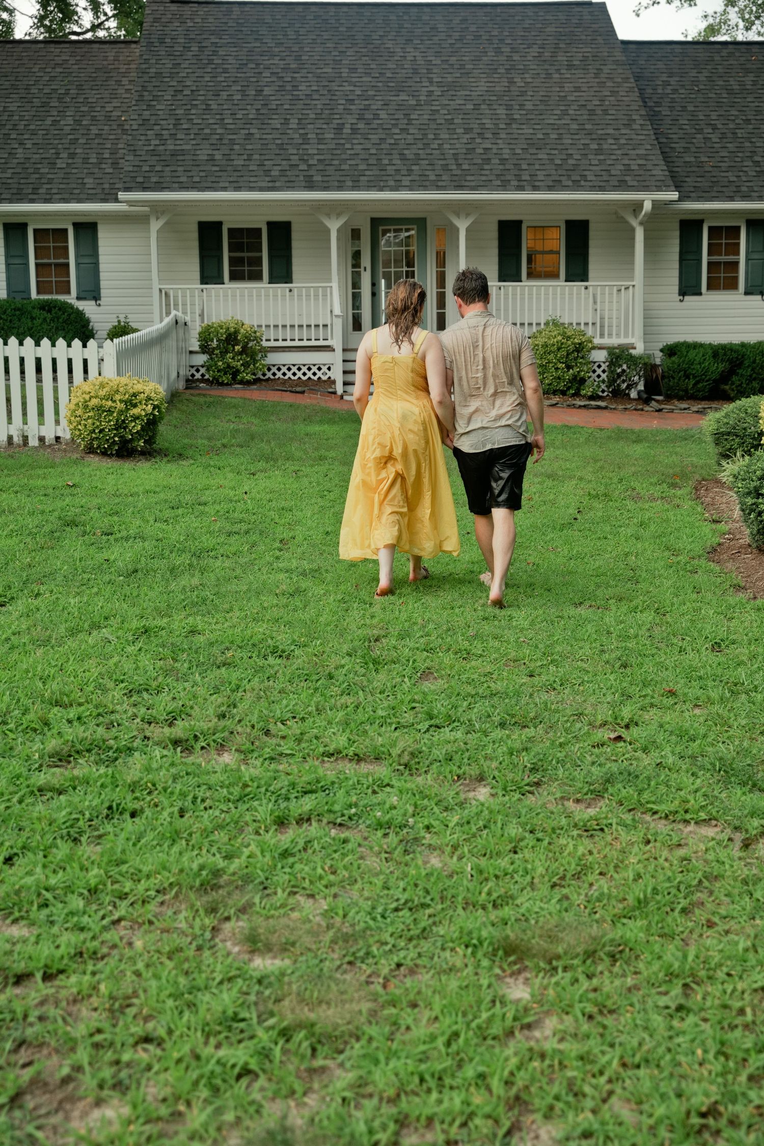 Couple walking hand-in-hand across green lawn toward charming white farmhouse with dark shutters and covered porch.