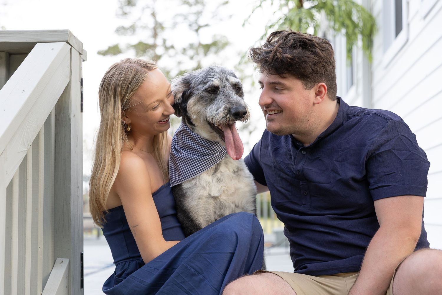 Smiling couple sitting on porch steps with fluffy gray dog wearing bandana.