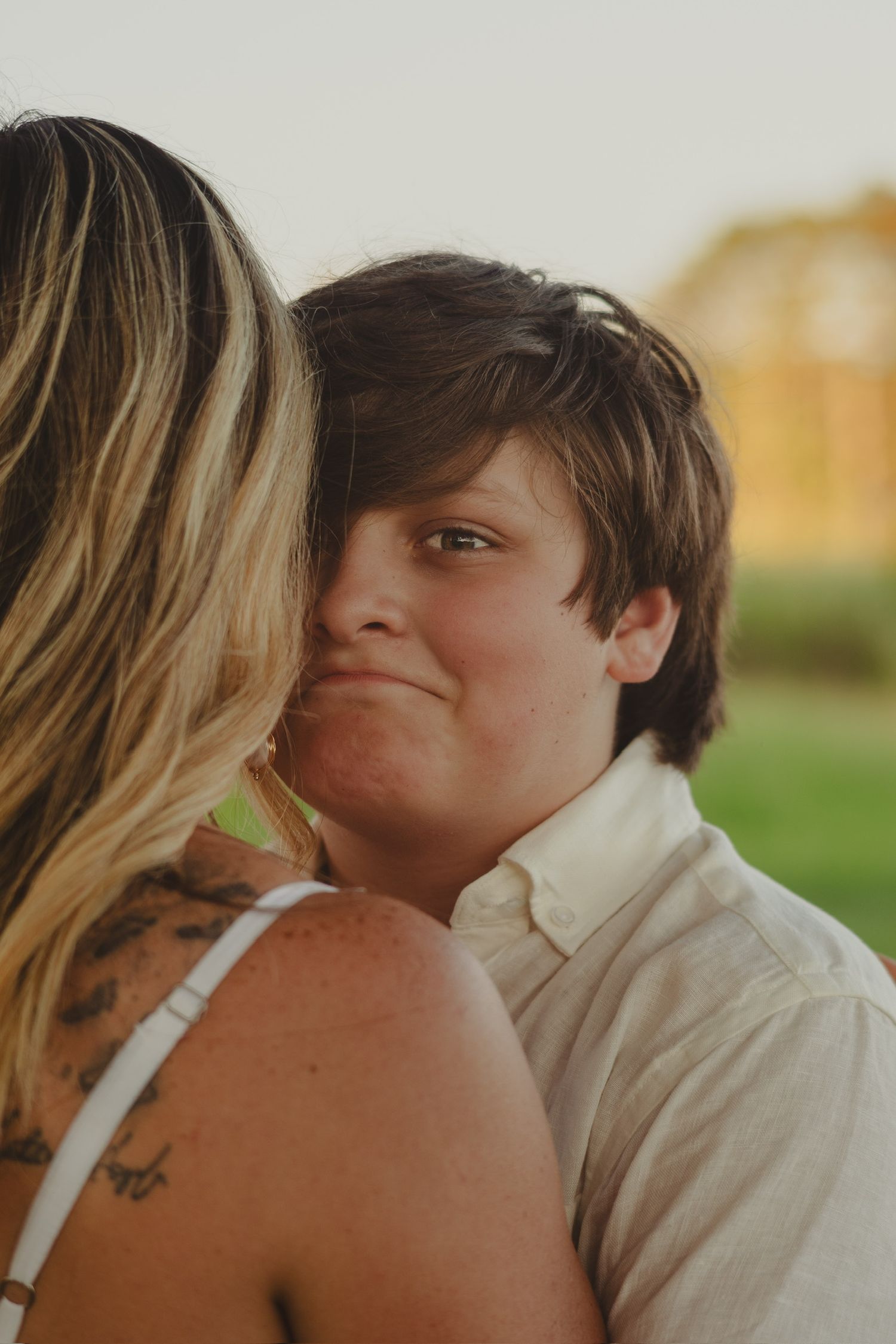 A young boy smiles warmly while being embraced by a tattooed woman outdoors in soft golden light.
