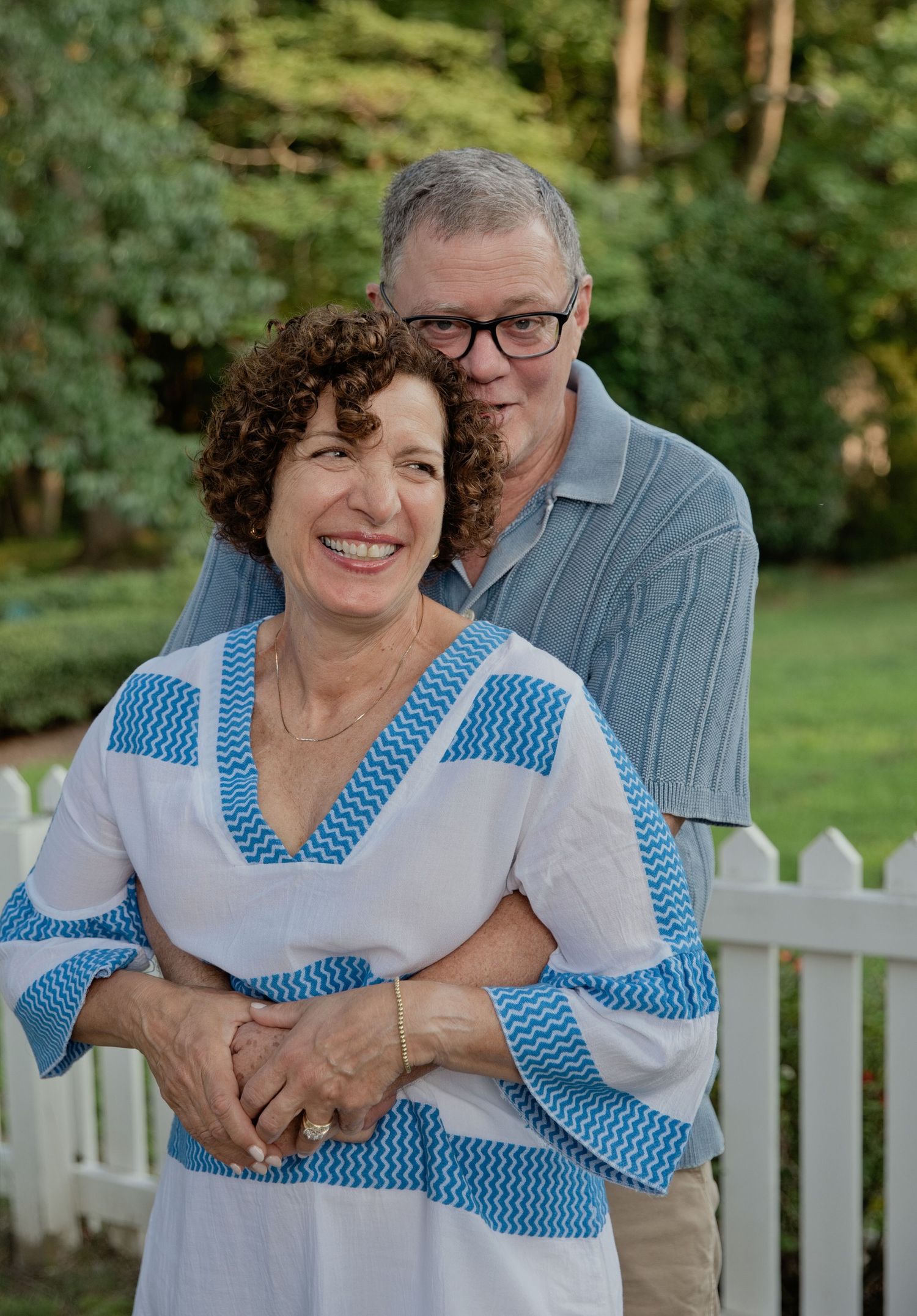 Smiling older couple posing outdoors near white picket fence, woman in blue and white top, man in gray polo shirt.