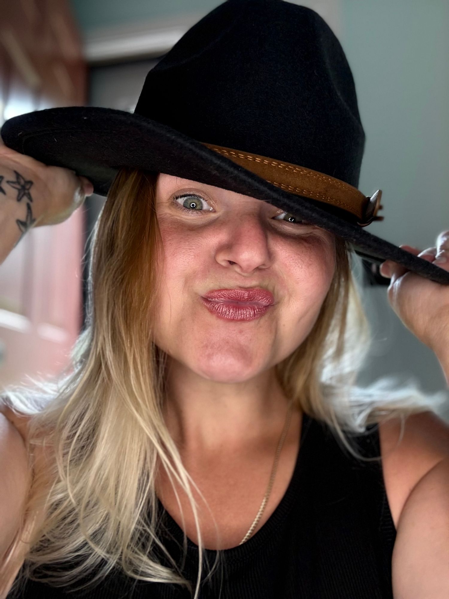 Blonde woman making a pouty face while wearing a black wide-brim fedora hat with brown band.