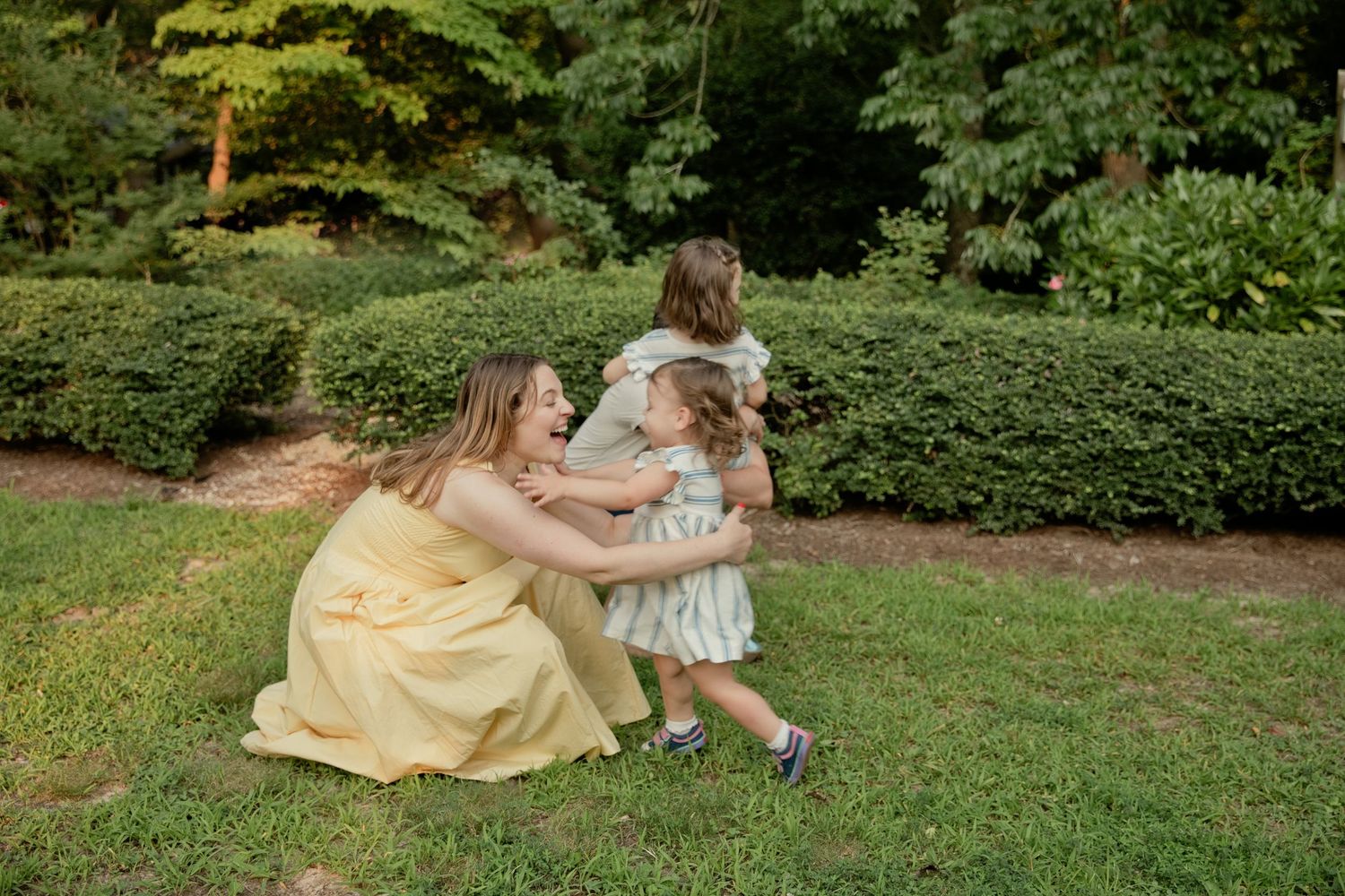 A loving mother in a yellow dress kneels on grass hugging her two small children in a garden.