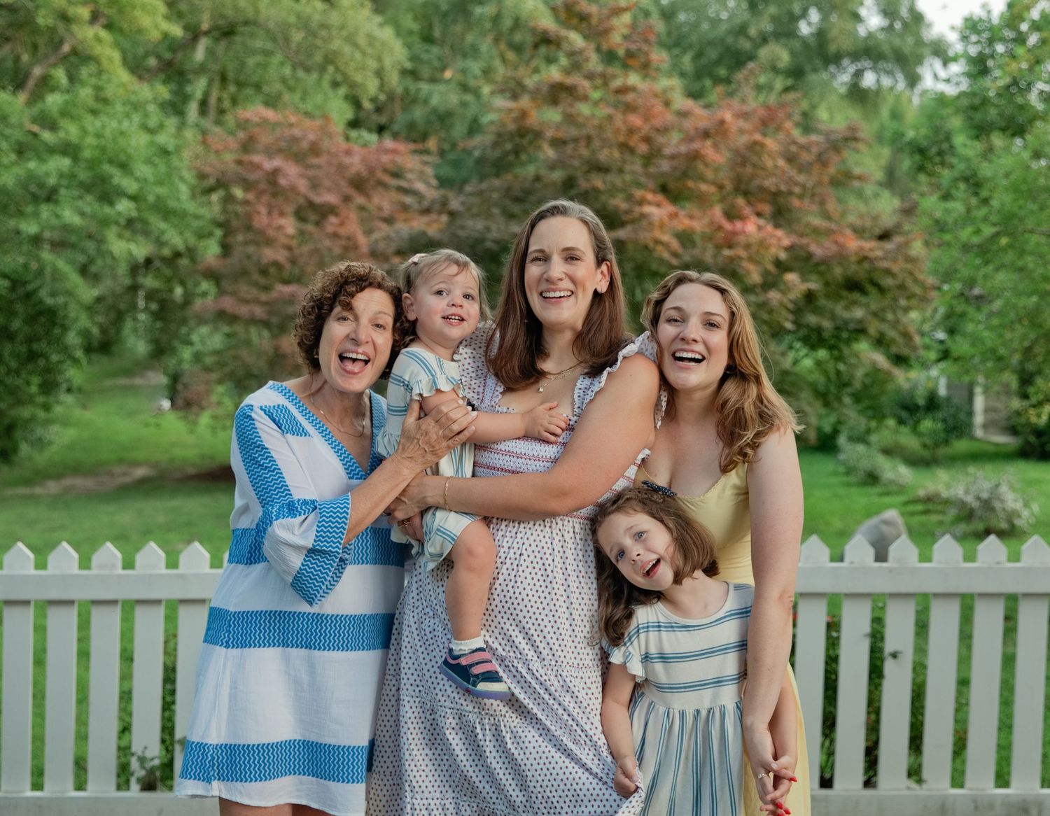 Three generations of women smile together by a white fence, holding two young children outdoors.