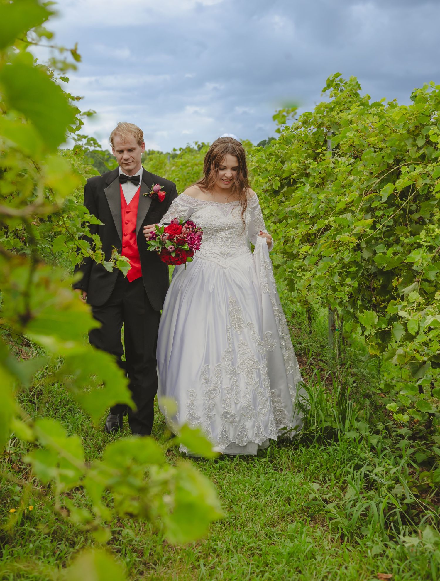 Bride and groom walking through lush Zoll's vineyard, VA, bride in white lace gown holding red bouquet, groom in black tuxedo with red vest.