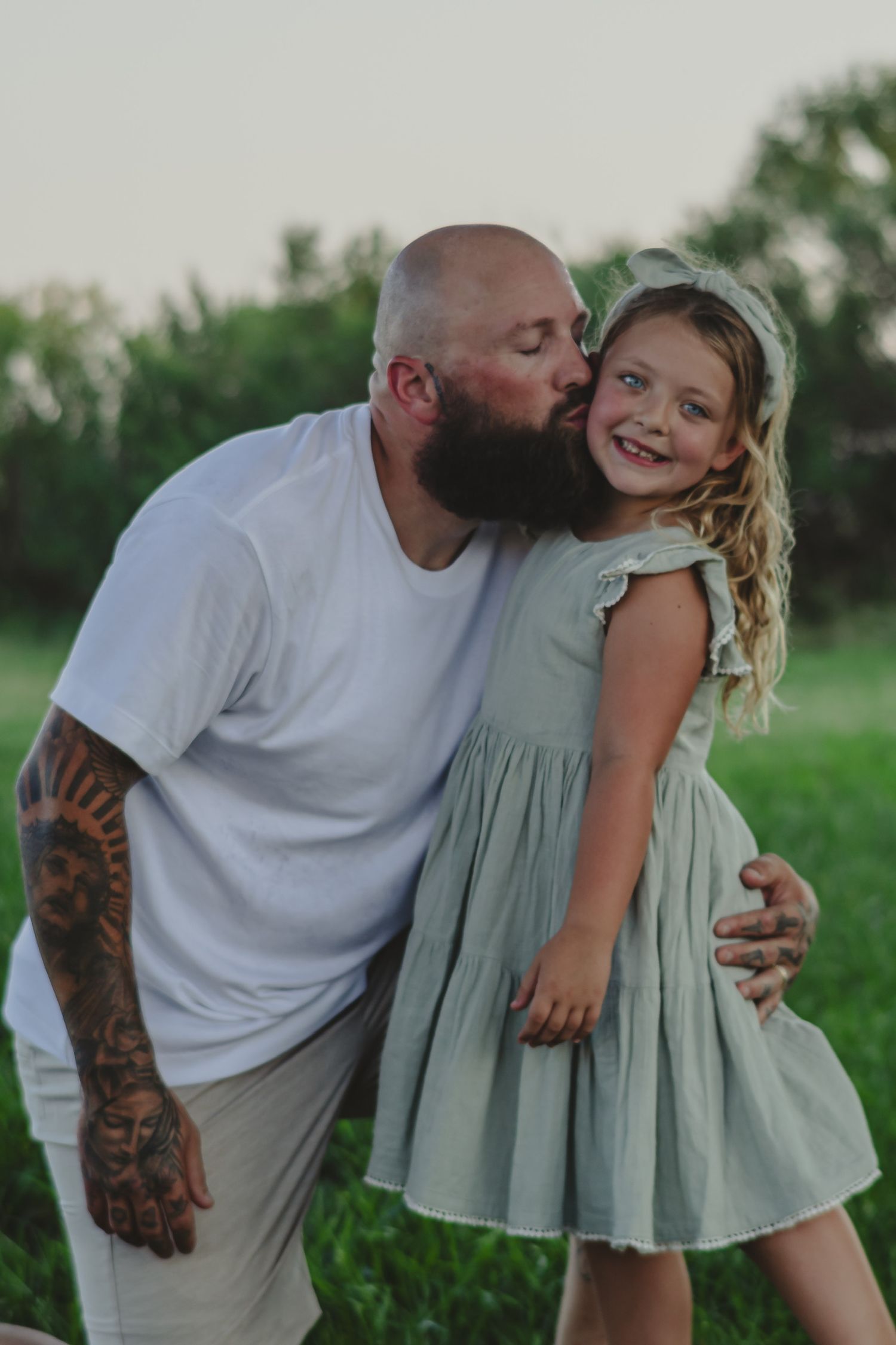 Tattooed bald man with beard kissing smiling little girl in sage green dress outdoors on green field.