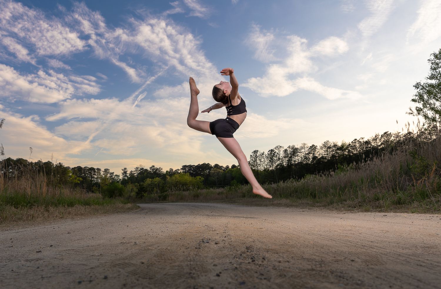 A dancer leaps gracefully against a blue sky, performing a stunning jump on an outdoor sandy path surrounded by trees.