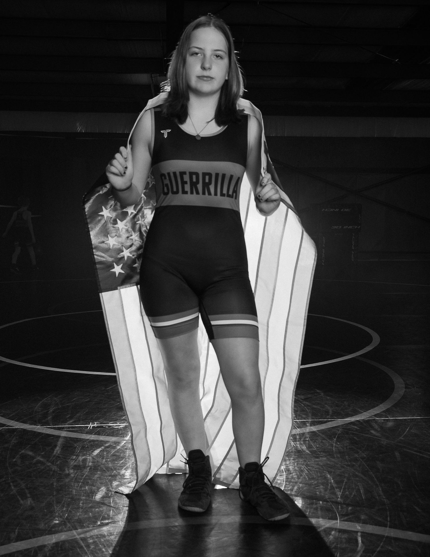 Smiling young female wrestler in black and pink singlet wearing a tiara poses on a wrestling mat with an American flag in background.