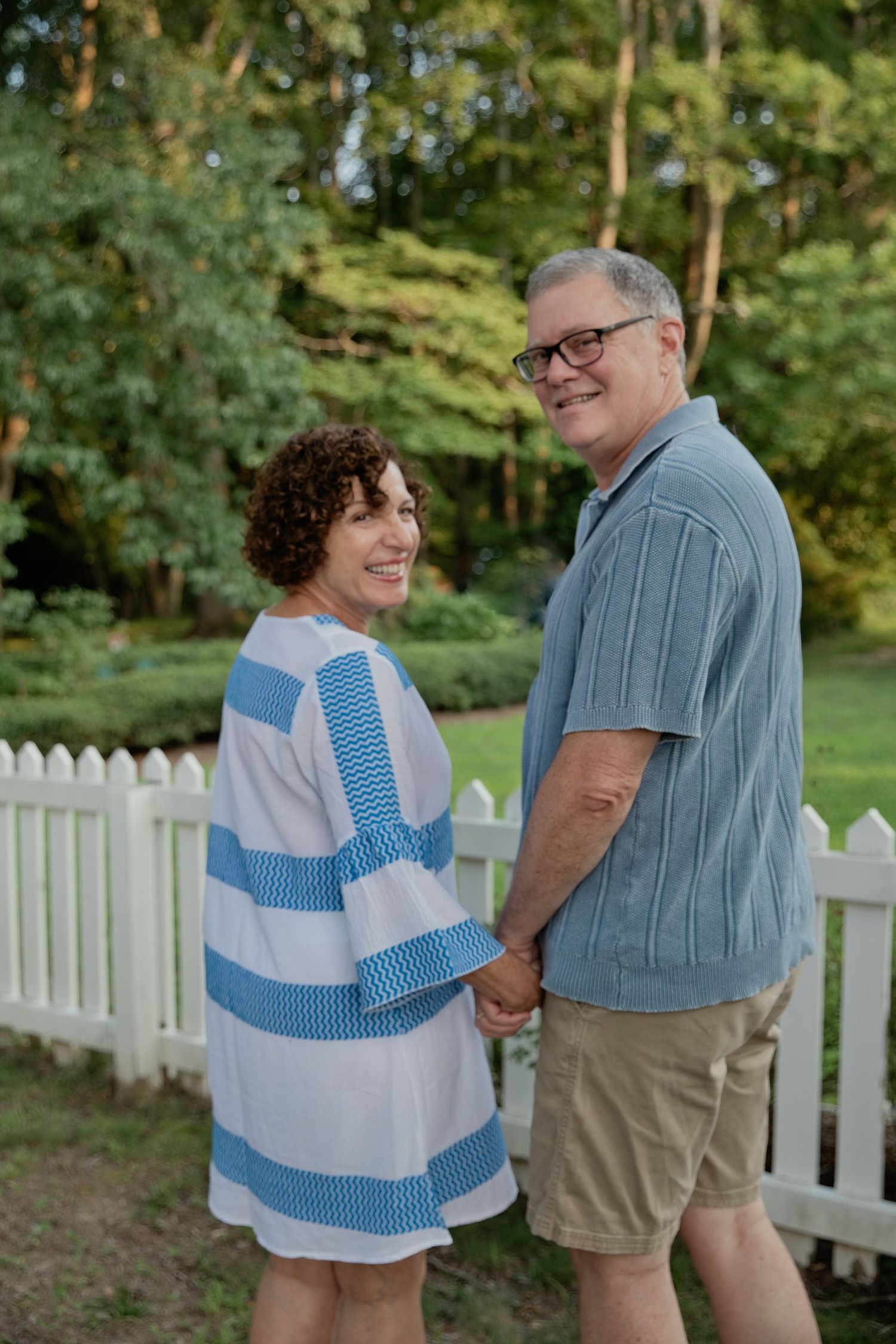 A smiling middle-aged couple holds hands in a lush backyard garden near a white picket fence.