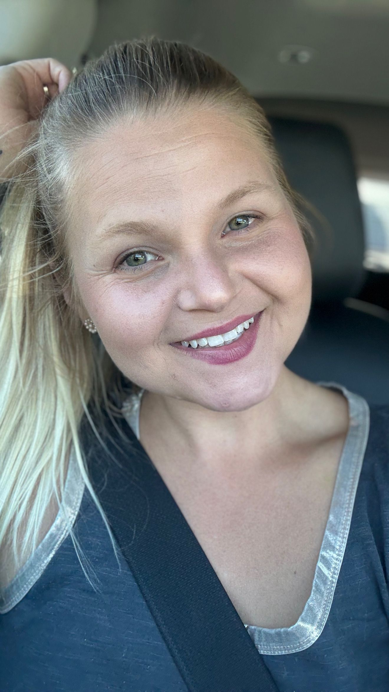 Smiling blonde woman taking a selfie in a car, wearing a gray top and seatbelt, with natural makeup.