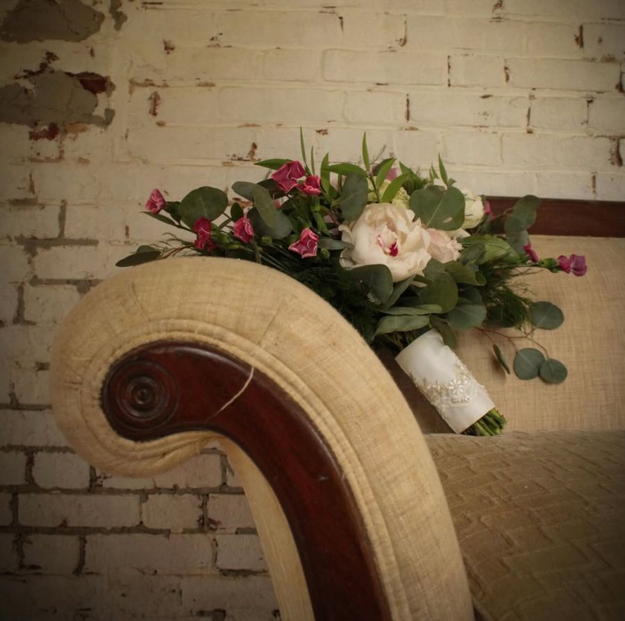 Bridal bouquet with white roses, pink flowers, and eucalyptus resting on a vintage wooden chair against a white brick wall.